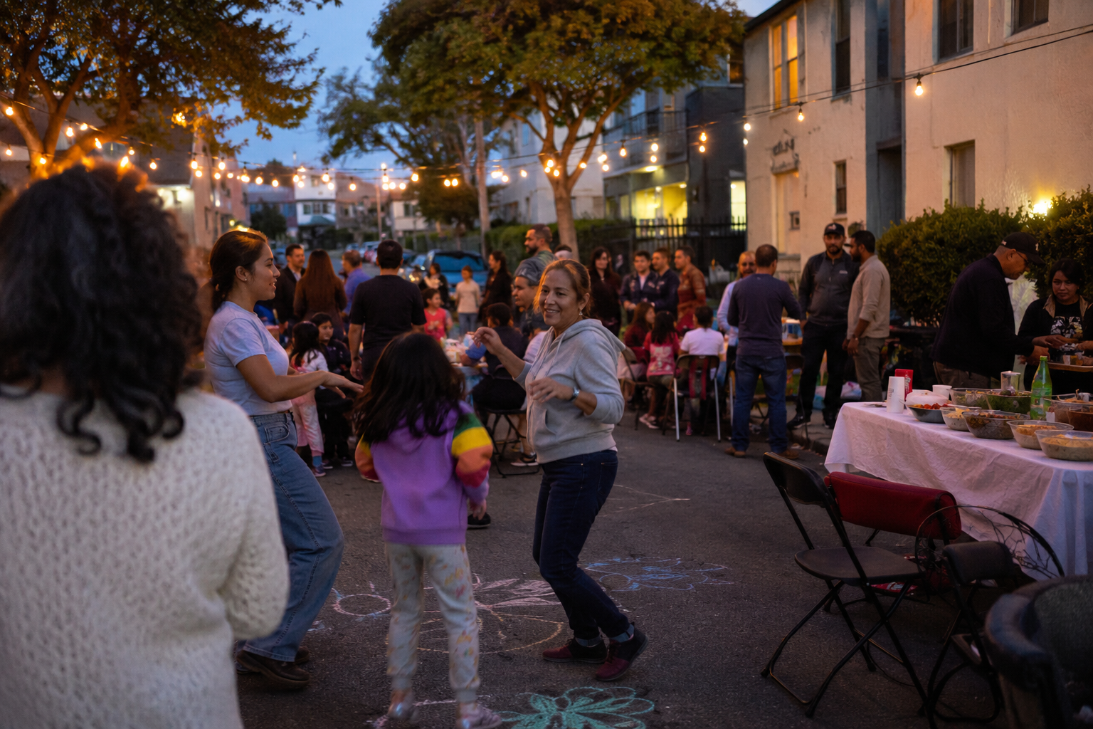 Neighbors eating, talking and dancing at a small Los Angeles block party under string lights.
